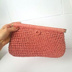 May include: A coral-colored woven clutch with a fold-over flap. The clutch features a textured, basket-weave design and a matching coral-colored flap with a button closure. Gold-toned hardware accents the flap's edges.
