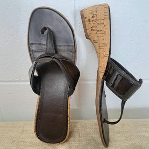 Puede incluir: Un par de sandalias de cuero marrón con cuñas de corcho. Las sandalias presentan un diseño de estilo chancla con una correa en el tobillo. La plantilla es de color marrón oscuro y la cuña es de corcho.