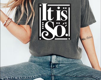 May include: Dark grey t-shirt featuring the white text "It is So". The t-shirt is worn with blue jeans and white slip-on shoes. The outfit has a relaxed, casual style.