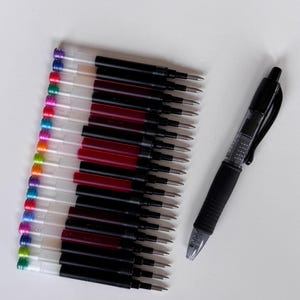 May include: A collection of black pen refills with various colored tops, alongside a black and clear pen. The refills are arranged in rows, showcasing a range of ink colors. The pen has a black grip and a clear barrel, providing a view of the ink.
