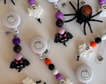 May include: Halloween-themed retractable badge holders with white pumpkin designs, beaded accents, and attached spider, ghost, and bat charms. The spiders have glittery orange backs. The beads are black, white, orange, and purple.