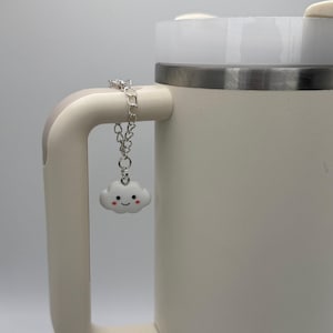 May include: A white ceramic mug with a silver chain attached to the handle. The chain has a small white cloud charm with a pink smiling face.