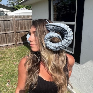 May include: A woman with long blonde hair wearing a black tank top and blue jeans. She is wearing a pair of large, grey, curled horns on her head.