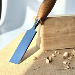 May include: A close-up of a wood carving chisel with a wooden handle. The handle is engraved with the word "SIRYI". The chisel is resting on a piece of wood with wood shavings around it.