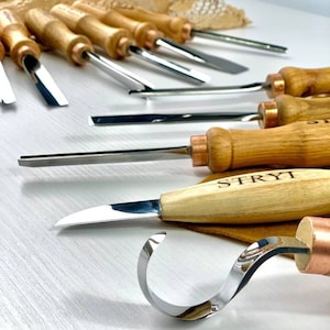 May include: A set of wood carving tools with wooden handles and metal blades. The tools include a hook knife, a gouge, and a straight chisel. The tools are arranged on a white surface. The tools are labelled "STRYI".