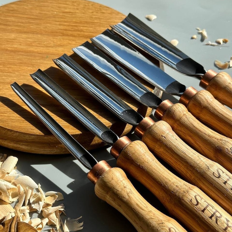 Pu&ograve; includere: Set di sette strumenti per intagliare il legno con ferrule in rame e manici in legno. Gli strumenti hanno lame di forme diverse e sono disposti su una superficie in legno.