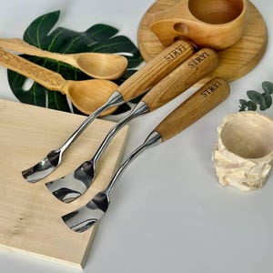 May include: A collection of wooden carving tools with stainless steel blades and wooden handles, alongside wooden spoons and a wooden cup. The handles of the tools are engraved with the word "STRYI". The items are arranged on a light-colored surface.