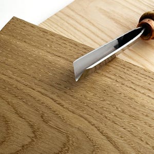 Op de afbeelding: Een houtbeitel met een gepolijst stalen lemmet en een houten handvat ligt op twee houten planken. De planken hebben verschillende bruintinten, met zichtbare houtnerf. Het lemmet van de beitel is schuin.