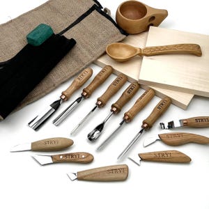 May include: A collection of wood carving tools with wooden handles and metal blades, including gouges, knives, and a spoon. A wooden cup and spoon are also present, along with a green block and a black bag. The tools are labeled with the word "STRYI".