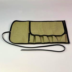 May include: A green canvas tool roll with black trim and a black strap. The roll is open, revealing several pockets. The canvas has a textured appearance. The strap is thin and appears to be made of leather or a similar material.
