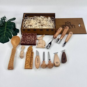 May include: A selection of wooden carving tools and decorative wooden items, such as a spoon, a rabbit figurine, and a carved block. The tools feature wooden handles and metal blades. The set is presented with a wooden box filled with straw.