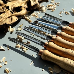 Pu&ograve; includere: Un set di sei strumenti per intagliare il legno con manici in legno e dettagli in rame. Gli strumenti hanno diverse forme e dimensioni delle lame. Gli strumenti sono disposti su una superficie grigia con trucioli di legno.