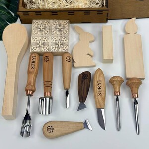May include: A selection of wooden carving tools and decorative wooden shapes, including a spoon, a bunny, and a candle. The tools have wooden handles and metal blades. A wooden box filled with wood shavings is in the background.