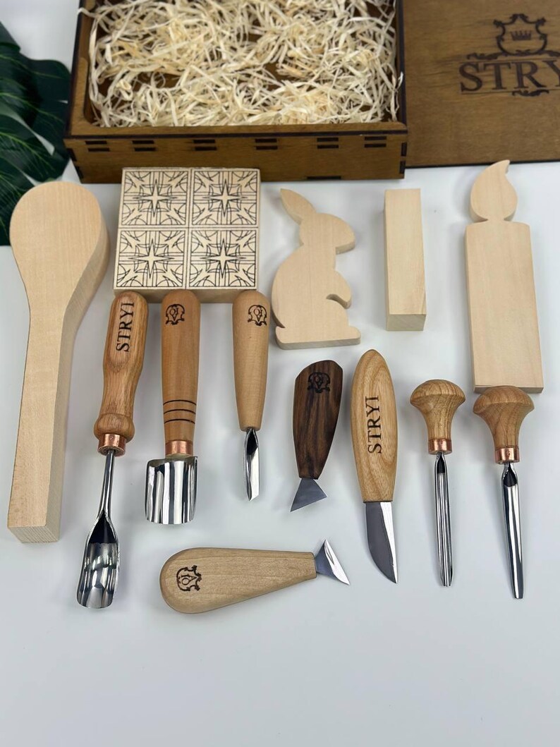 May include: A selection of wooden carving tools and decorative wooden shapes, including a spoon, a bunny, and a candle. The tools have wooden handles and metal blades. A wooden box filled with wood shavings is in the background.