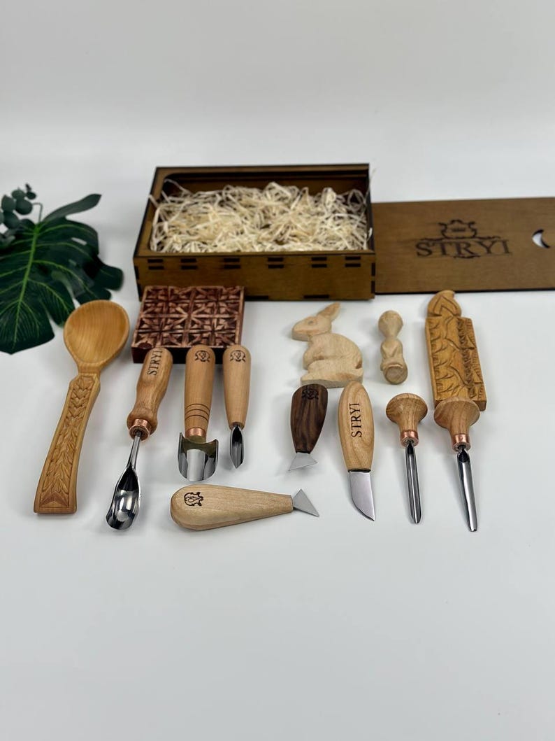 May include: A selection of wood carving tools and a wooden box with the text "STRYI". The tools include chisels, knives, and a spoon, all with wooden handles and metal blades. A carved wooden rabbit and other decorative wooden pieces are also included.