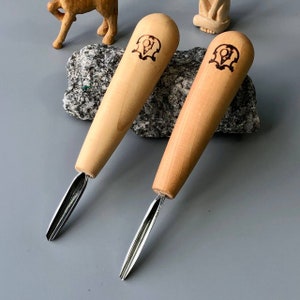 Puede incluir: Dos herramientas de talla de madera con cuchillas de metal. Las herramientas tienen un mango de madera marrón con un diseño quemado. Las herramientas descansan sobre una roca gris.