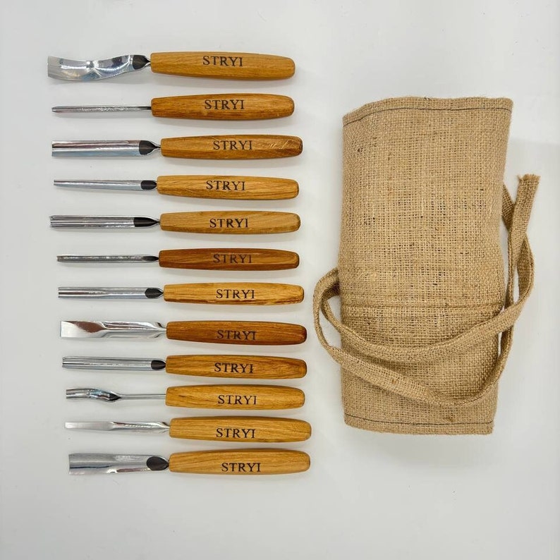 May include: A set of 12 wood carving tools with wooden handles and metal blades. The tools are arranged in a row on a white surface. The tools are labelled "STRYI" on the handles. The tools are packaged in a brown hessian bag.