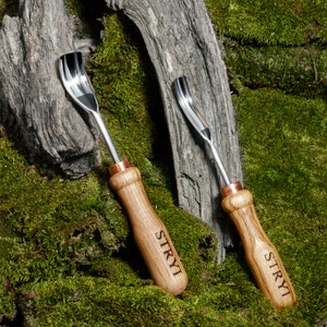 Pu&ograve; includere: Due strumenti per intagliare il legno con accenti in rame e la parola "STRYI" incisa sui manici. Gli strumenti sono appoggiati su una superficie ricoperta di muschio.