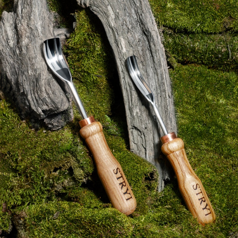 Pu&ograve; includere: Due strumenti per intagliare il legno con accenti in rame e la parola "STRYI" incisa sui manici. Gli strumenti sono appoggiati su una superficie ricoperta di muschio.