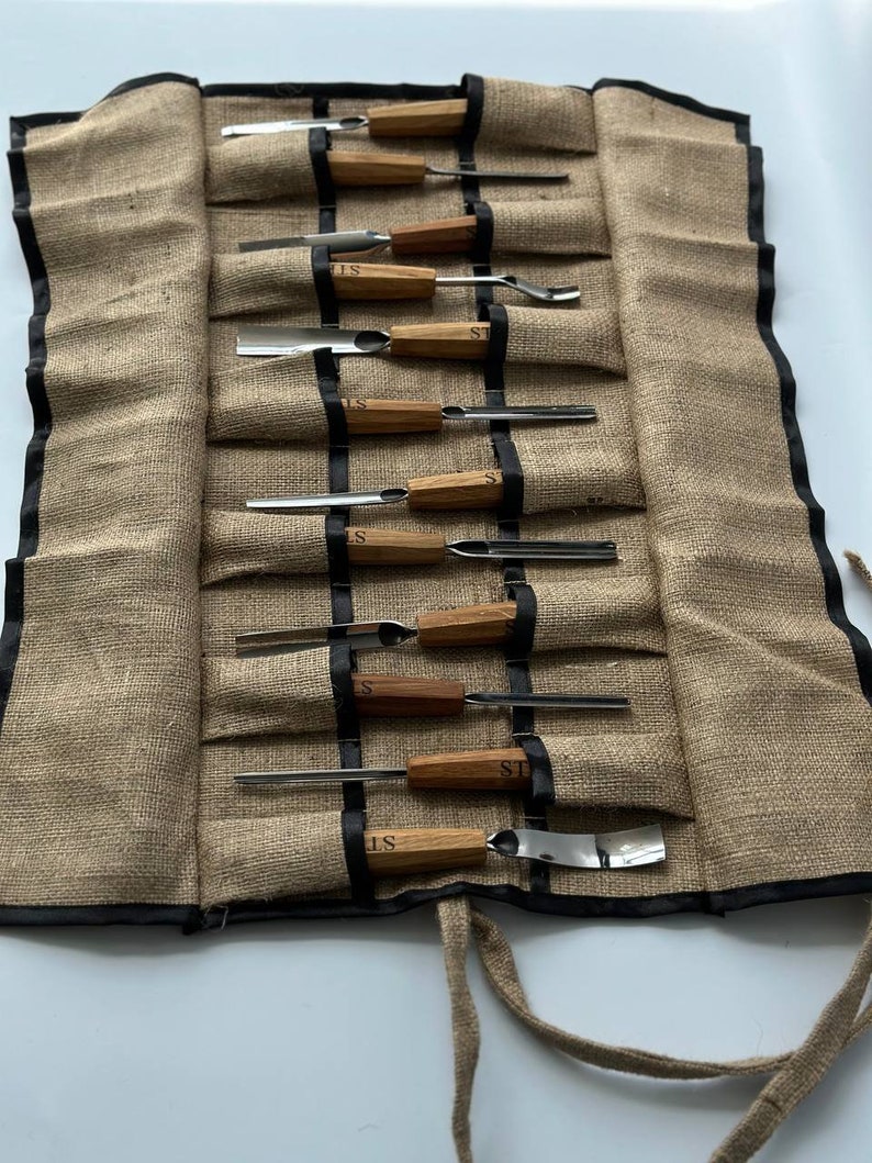 May include: A set of wood carving tools with wooden handles, rolled up in a brown hessian carrying case with black trim. The tools are arranged in rows and secured with black straps.