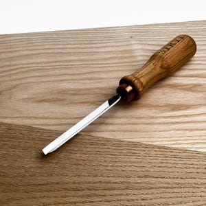 Peut inclure: Un ciseau à bois avec une lame en acier poli et un manche en bois. Le manche est gravé du mot "STRY". Le ciseau est posé sur une surface en bois brun clair.