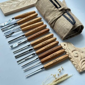May include: A set of 10 wood carving tools with wooden handles and metal blades. The tools are arranged in a row on a white surface. The tools are labelled "STRYI" on the handles. The tools are accompanied by a rolled-up hessian bag with a black ribbon, a piece of carved wood, and a sprig of dried flowers.