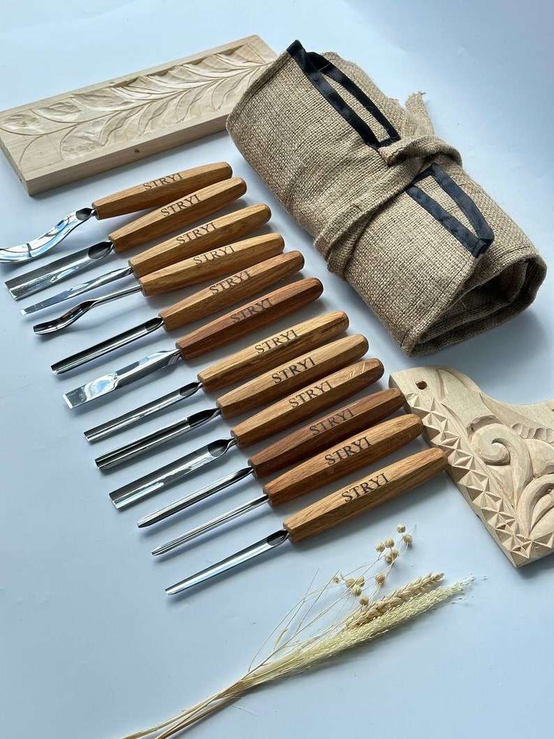 May include: A set of 10 wood carving tools with wooden handles and metal blades. The tools are arranged in a row on a white surface. The tools are labelled "STRYI" on the handles. The tools are accompanied by a rolled-up hessian bag with a black ribbon, a piece of carved wood, and a sprig of dried flowers.