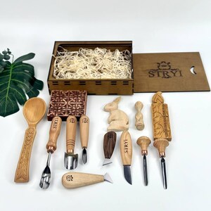 May include: A selection of wood carving tools and accessories, including chisels, knives, and a wooden spoon, displayed on a white surface. A wooden box with the word "STRYI" is also present, alongside a carved wooden rabbit and other decorative items.