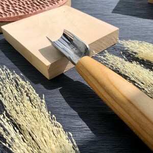 Puede incluir: Una herramienta de talla de madera con una cuchilla curva y un mango de madera. La herramienta está apoyada sobre una superficie de madera.