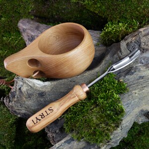 Pu&ograve; includere: Una tazza di legno con un manico e un piccolo foro sul lato, accanto a uno strumento di legno con una punta di metallo e la parola "STRYI" incisa sul manico.