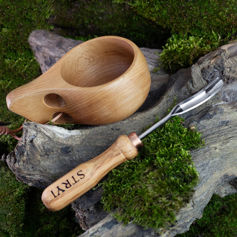 Pu&ograve; includere: Una tazza di legno con un manico e un piccolo foro sul lato, accanto a uno strumento di legno con una punta di metallo e la parola "STRYI" incisa sul manico.