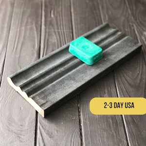 May include: A black concrete soap dish with three grooves to hold soap bars. A teal green soap bar is resting in the dish. The dish is on a dark wood surface. 2-3 Day USA.