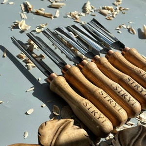 Pu&ograve; includere: Un set di otto strumenti per intagliare il legno con manici in legno. I manici sono incisi con la parola "STRYI". Gli strumenti sono disposti su una superficie grigia con trucioli di legno.