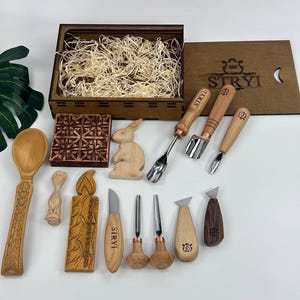 May include: A selection of wood carving tools and carved wooden objects. The set includes knives, gouges, and spoons, along with a wooden box. The box lid is branded with "STRYI". Carved items include a rabbit, candle, and decorative block.
