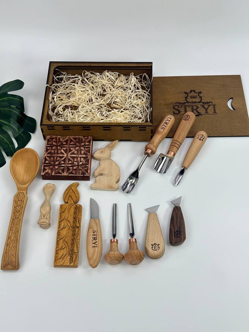 May include: A selection of wood carving tools and carved wooden objects. The set includes knives, gouges, and spoons, along with a wooden box. The box lid is branded with "STRYI". Carved items include a rabbit, candle, and decorative block.