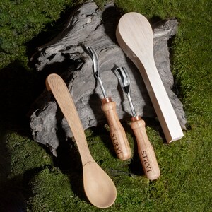 Pu&ograve; includere: Quattro utensili in legno, tra cui un cucchiaio, due strumenti da intaglio e una pala di legno. Gli strumenti da intaglio hanno manici in rame e il testo "STRYI" inciso su di essi. Gli utensili sono su una superficie verde muschiosa.