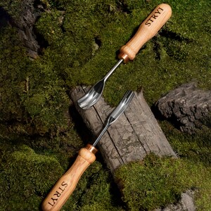 Pu&ograve; includere: Due strumenti di intaglio in legno con lame in metallo. Gli strumenti hanno un manico in legno marrone con la parola "STRYI" incisa su di essi. Gli strumenti sono su un letto di muschio verde.