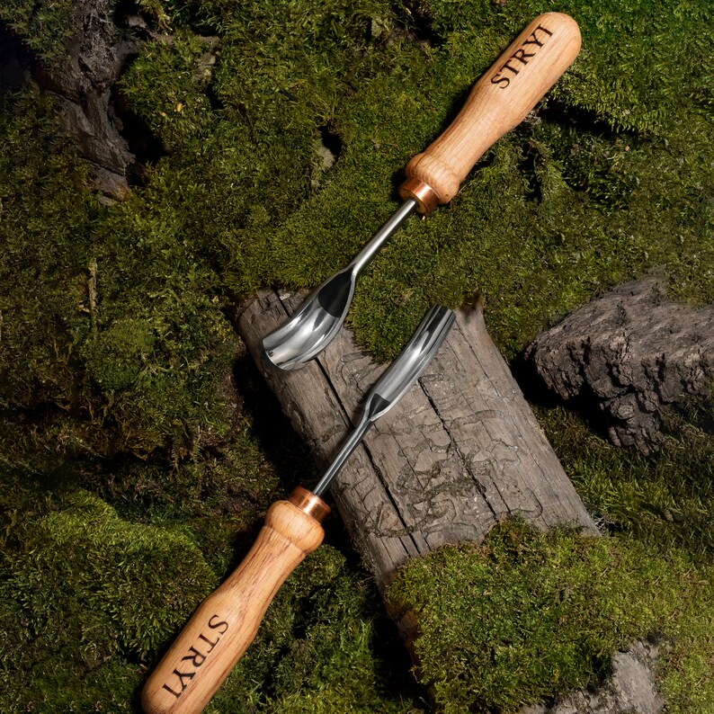 Pu&ograve; includere: Due strumenti di intaglio in legno con lame in metallo. Gli strumenti hanno un manico in legno marrone con la parola "STRYI" incisa su di essi. Gli strumenti sono su un letto di muschio verde.