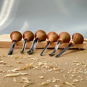 May include: A set of six wood carving tools with wooden handles and metal blades. The tools are arranged on a wooden surface with wood shavings scattered around them.