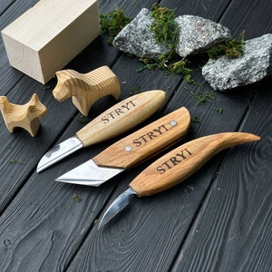 May include: Three wood carving knives with wooden handles and stainless steel blades, each engraved with "STRYI". Two small wooden animal figurines and a rectangular wooden block are also visible. The items are arranged on a dark wooden surface.