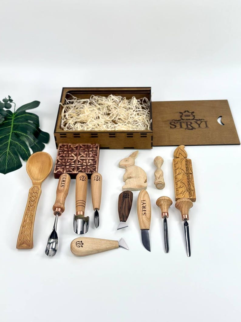 May include: A selection of wood carving tools and a wooden box filled with wood shavings. The tools include chisels, knives, and a spoon, all with wooden handles. The box lid has the word "STRYI" printed on it.