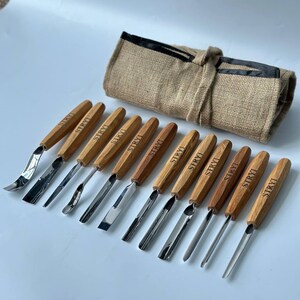 May include: A set of 10 wood carving tools with wooden handles and metal blades. The tools are arranged in a row on a white surface. The tools are all labelled "STRYI". The tools are rolled up in a brown hessian bag with black trim.