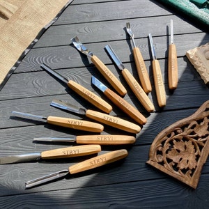 Puede incluir: Un conjunto de 11 herramientas de talla de madera con mangos de madera. Las herramientas tienen diferentes formas y tamaños para tallar madera. Las herramientas están dispuestas en forma de abanico sobre una superficie negra. Las herramientas están etiquetadas con "STRYI".