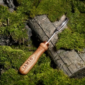 Pu&ograve; includere: Uno strumento con manico in legno e denti in metallo. Lo strumento &egrave; inciso con la parola "STRYT". Lo strumento &egrave; appoggiato su un letto di muschio verde.