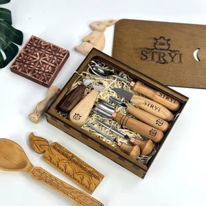 May include: A wooden carving set with various tools, including knives and gouges, presented in a wooden box. The box is branded with the text "STRYI". Additional wooden carvings, such as a spoon and a bunny, are also visible.