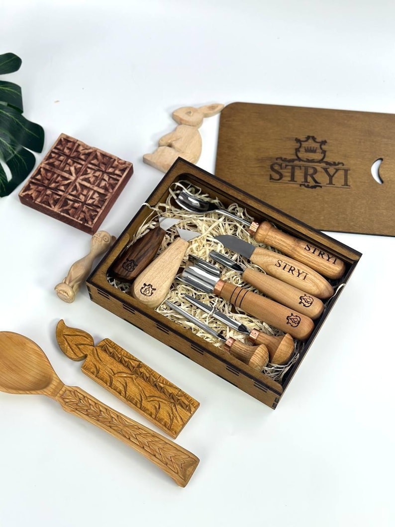 May include: A wooden carving set with various tools, including knives and gouges, presented in a wooden box. The box is branded with the text "STRYI". Additional wooden carvings, such as a spoon and a bunny, are also visible.