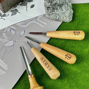 May include: Four wood carving tools with wooden handles and metal blades. The tools are arranged on a green surface with a piece of paper and two gray rocks in the background.