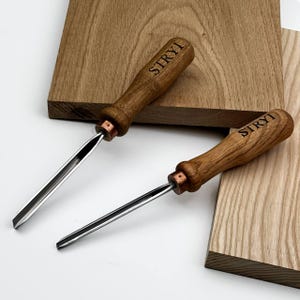 May include: Two wood carving tools with the word "STRYI" engraved on the wooden handles. The tools have a metal blade and copper-colored ferrule. The tools are resting on a wooden surface.