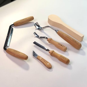 Puede incluir: Un juego de seis herramientas de talla de madera con mangos de madera. Las herramientas tienen cuchillas de metal y están etiquetadas como "STRYI". El juego también incluye un blanco de cuchara de madera.