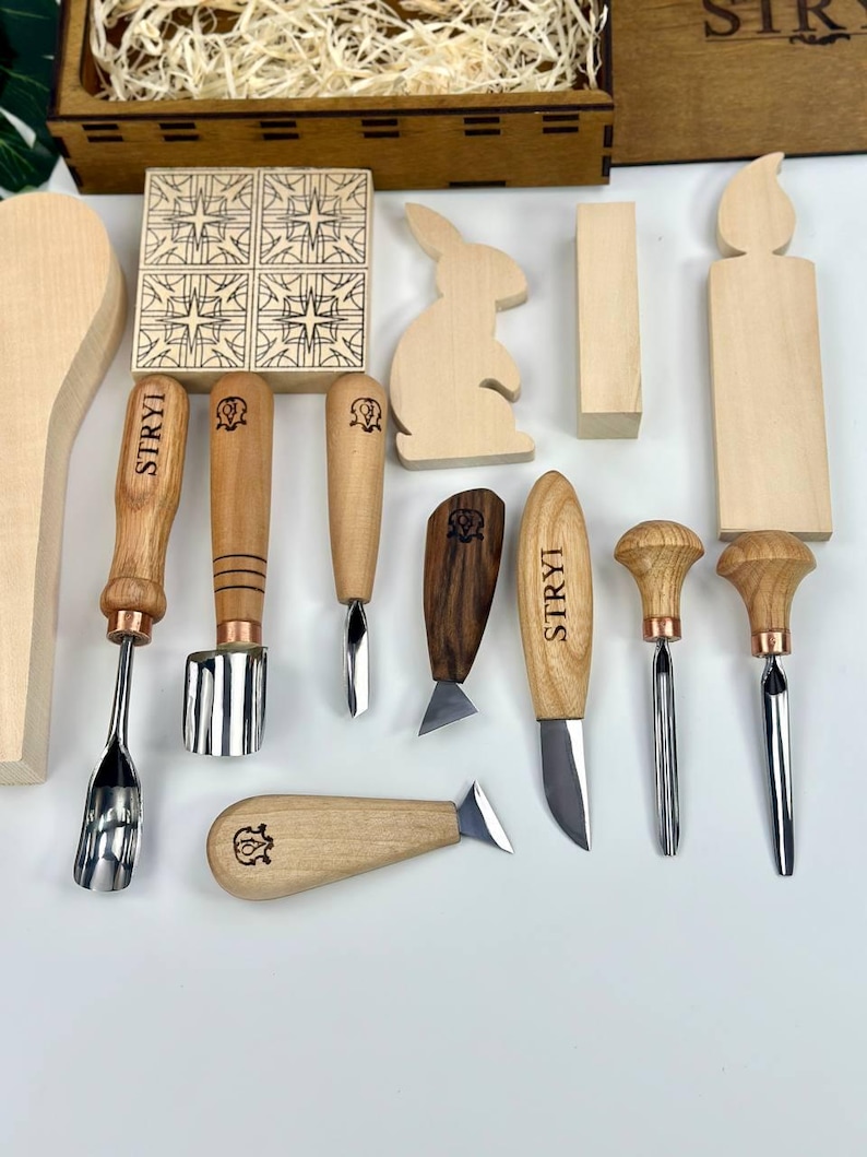 May include: A selection of wood carving tools and wooden shapes, including a rabbit, candle, and geometric tiles. The tools have wooden handles and metal blades. A wooden box with the word "STRYI" is in the background.